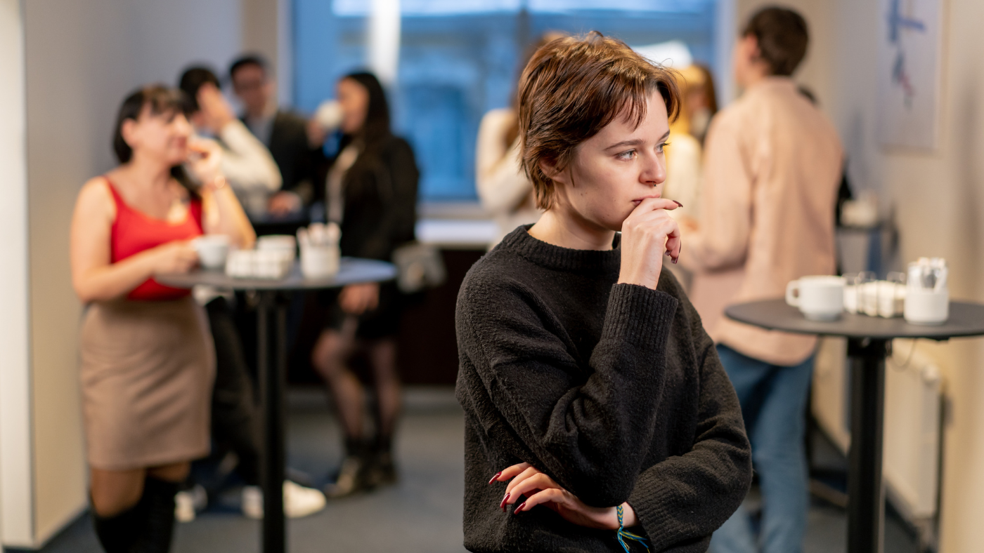 A young woman stands alone with her arms crossed at a busy Berlin social gathering while others chat in the background — feeling like she has no friends in Berlin