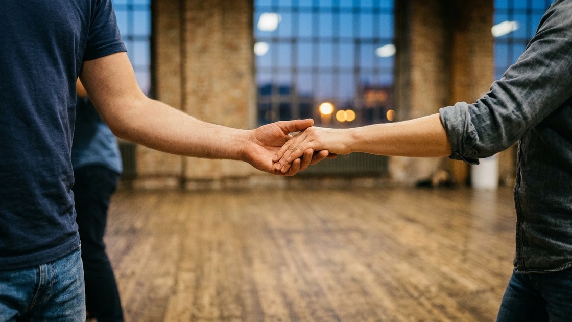 Two dancers' joined hands in a simple beginner hold, with a worn wooden studio floor blurred behind them