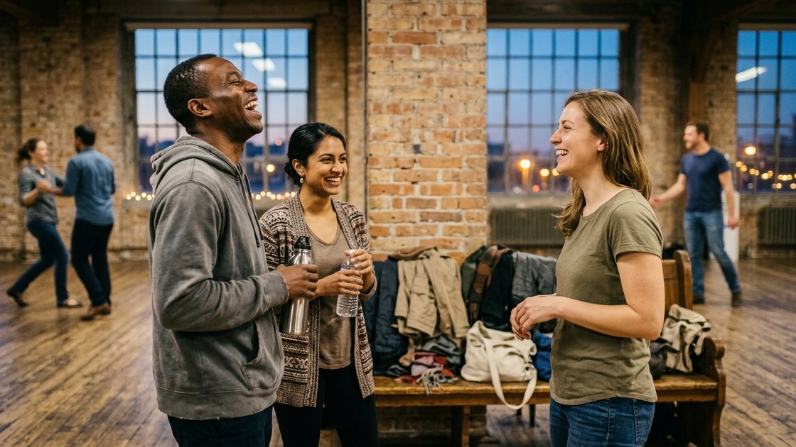 Three people laughing and drinking water at the edge of a dance studio between songs, coats piled on a bench