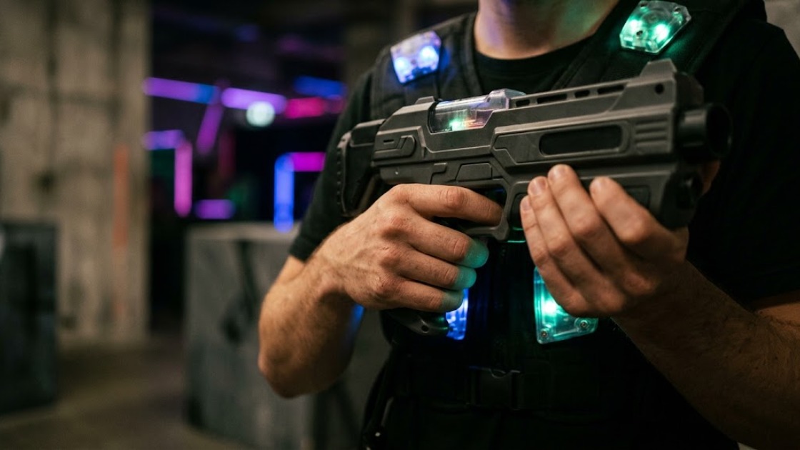 A player's hands gripping a laser tag phaser, glowing vest sensors behind, in a dark neon-lit Berlin arena