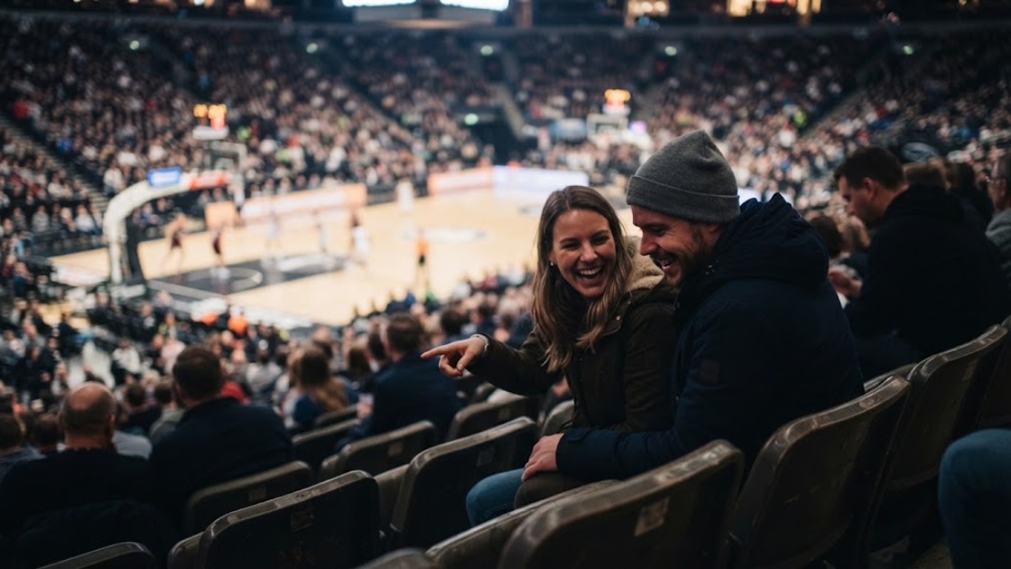 Two friends laughing in the upper seats of a packed basketball arena during a game