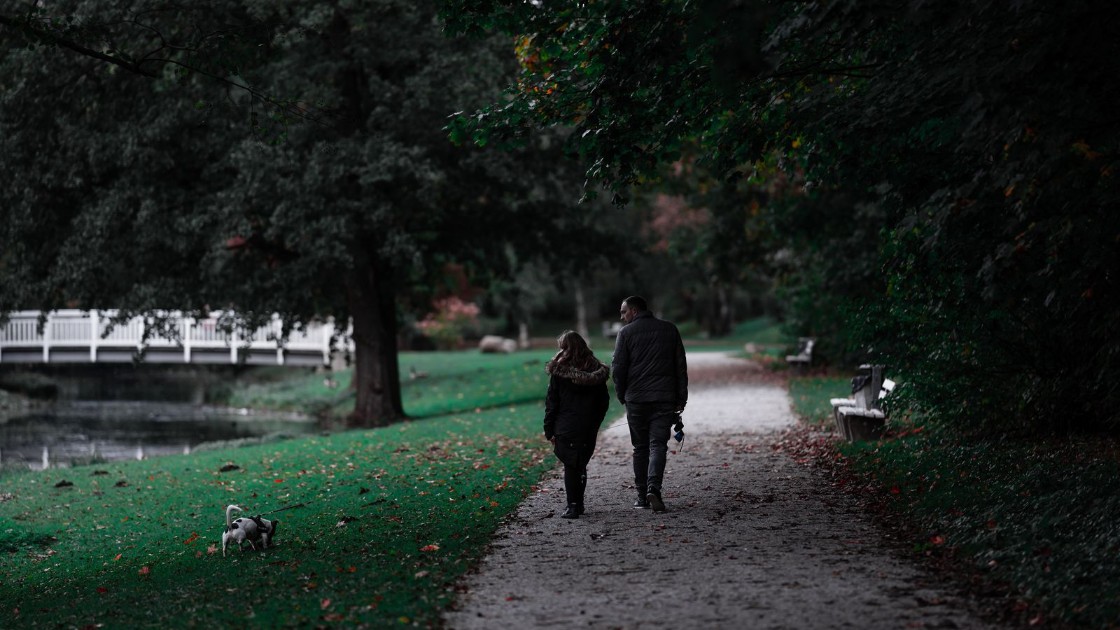 Two people walking a small dog along a leafy park path in autumn