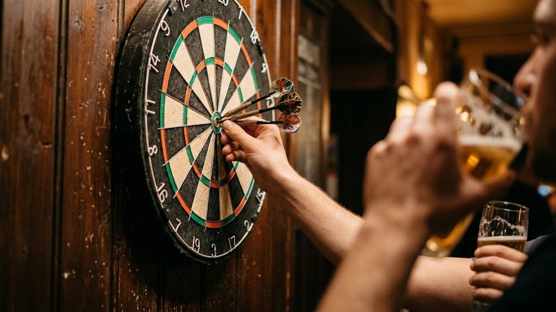 A hand pulls three darts out of a bristle dart board while a friend raises a beer in the foreground