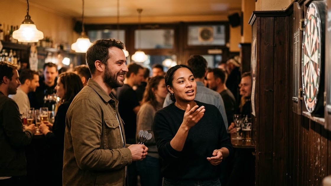 Two players chat at the throw line in a busy Berlin bar at night, one holding three darts