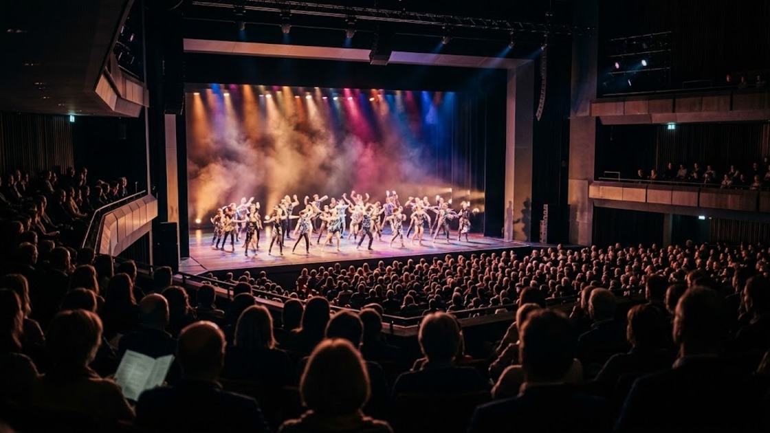 View from the high stalls of a large theatre over the audience towards dancers in glittering costumes on a lit stage