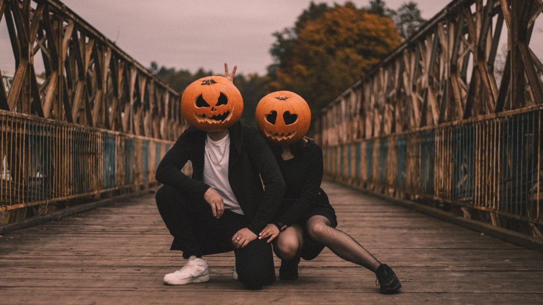 Couple in black costumes with carved pumpkin heads crouching on an old wooden bridge