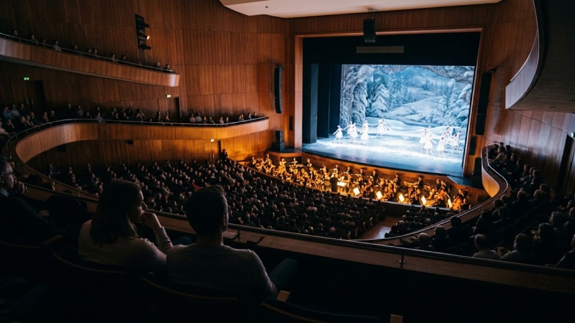 Two people watch a winter ballet from the upper seats of a large opera auditorium with a live orchestra