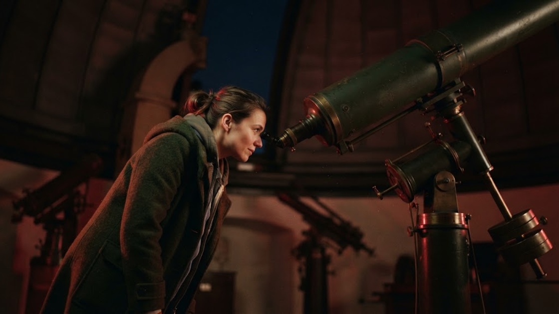 A woman in a winter coat bends towards the eyepiece of a long historic refracting telescope inside an observatory dome at night